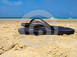 Black flip flops on a sand with blue sea and sky background