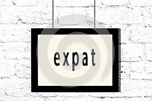 Black frame hanging on white brick wall with inscription expat
