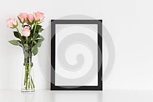 Black frame mockup with a bouquet of pink roses in a glass vase on a white table. Portrait orientation