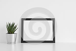 Black frame mockup with a cactus in a pot on a white table.Landscape orientation