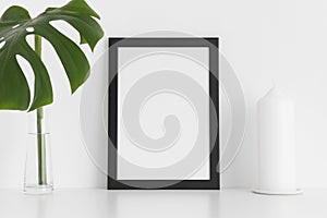 Black frame mockup with a candle and a monstera leaf in a vase on a white table.Portrait orientation