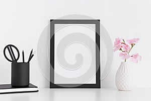 Black frame mockup with pink oleander in a glass vase and workspace accessories on a white table.Portrait orientation