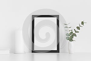 Black frame mockup with workspace accessories and eucalyptus in a vase on a white table. Portrait orientation