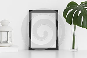 Black frame mockup with workspace accessories and a monstera leaf in a vase on a white table.Portrait orientation