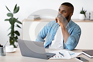 Black Freelancer Guy Sitting At Workplace With Thoughtful Face Expression
