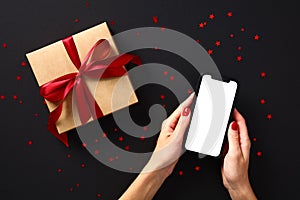 Black Friday online shopping concept. Mobile phone with blank screen mockup in female hands and gift box wrapped red ribbon over