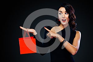 Black friday sale concept for shop. Shopping girl holding red bag isolated on dark background. Woman pointing to looking