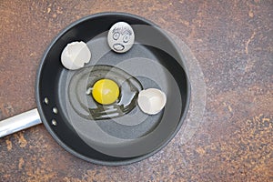 Black frying pan with raw broken egg and eggshell with emotion drawing on a rusty background