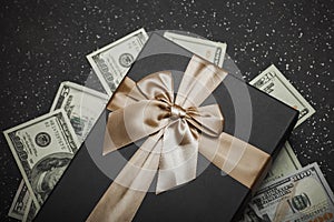 Black gift box with gold ribbon and a big bow on a pile of money. Gift on a granite surface