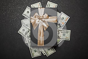 Black gift box with gold ribbon and a big bow on a pile of money. Gift on a granite surface