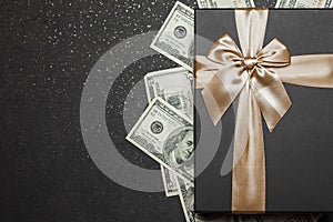 Black gift box with gold ribbon and a big bow on a pile of money. Gift on a granite surface