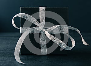 Black gift box with silver ribbon bow on black background. Tonned photo.