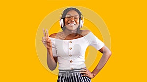 Black Girl In Headphones Gesturing V-Sign Standing Over Yellow Background