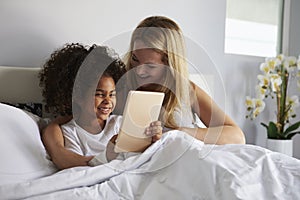 Black girl holds tablet computer, in bed with caucasian mum