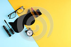 Black glasses, alarm clock, mascara, pomade and sleep mask on blue and yellow background composition