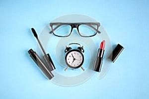 Black glasses, alarm clock, mascara and red pomade on blue background composition