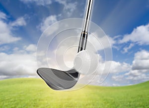 Black Golf Club Wedge Iron Hitting Golf Ball Against Grass and Blue Sky Background