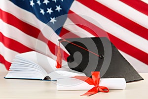 Black graduation hat, diploma and book on white wooden table against American flag