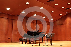 Black grand piano and bench with two chairs and music stands on light wood stage with medium wood walls and slanted ceiling lights