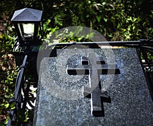 Black granite tombstone with an Orthodox cross on a grave at night in a cemetery with a luminous old lamp