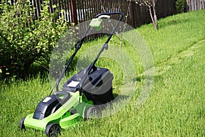 Black-green lawn mower stands on the lawn, with one strip of cut on the grass against the background of green bushes of