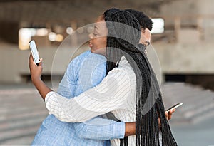 Black guy and girl hugging but looking at smartphones over shoulder