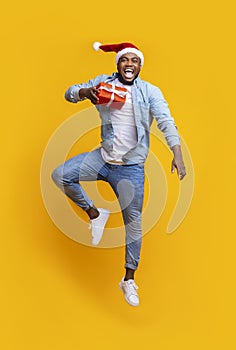 Black guy holding christmas gift and jumping over yellow background