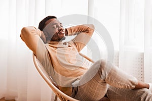 Black Guy Relaxing Sitting On Modern Chair Against Window Indoor