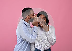 Black guy sharing secret or whispering gossips into his girlfriend's ear on pink studio background