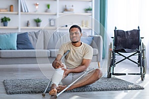 Black guy sitting on floor after falling with crutches, suffering from pain in broken leg, wheelchair standing nearby