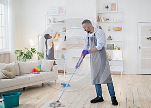 Black guy washing floor while his girlfriend cleaning shelves in their living room, empty space