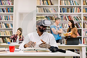 Black guy wearing vr goggles headset, reading book and using information from virtual reality, sitting in modern library