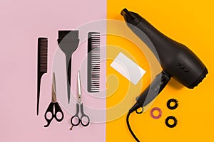 Black hair dryer, comb and scissors on pink and yellow paper background. Top view