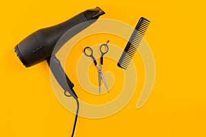 Black hair dryer, comb and scissors on yellow paper background. Top view