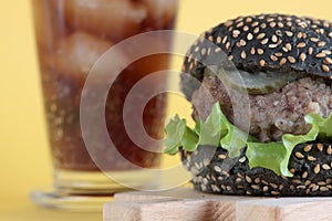Black hamburger on wooden cutting desk and a glass of ice cola  on yellow background. Unhealthy food concept