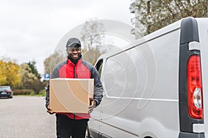 Black handsome delivery-person in red-and-gray jacket and black hat carrying one cardboard box. White delivery van