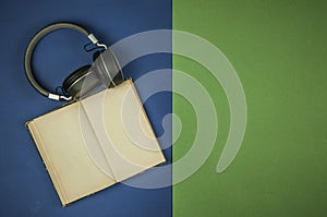 Black headphones with old open book with blank pages is lying on green and blue background. Copy space. Top view. Audio book
