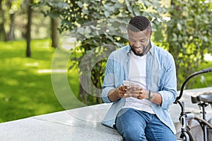 Black Hipster Guy Using Smartphone Outdoors, Sitting On Bench In Park