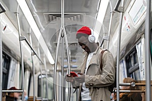 Black hipster man wear wireless headphones, listening to the music, using smartphone in subway train