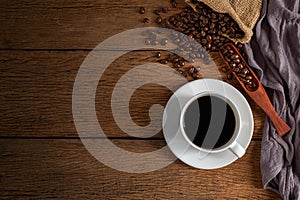 Black hot fresh coffee in a white ceramic cup with coffee beans roasted in burlap sack bag on wooden table background.