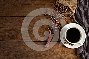 Black hot fresh coffee in a white ceramic cup with coffee beans roasted in burlap sack bag on wooden table background.