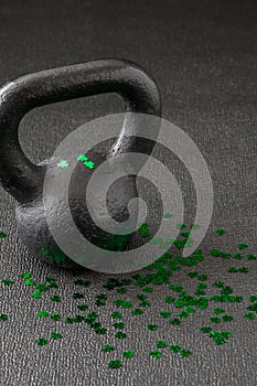 Black iron kettlebell covered in green shamrock confetti on a black gym floor, happy St. Patrick’s Day