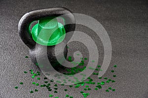 Black iron kettlebell, and green glitter leprechaun hat, covered in green shamrock confetti on a black gym floor, happy St. Patric