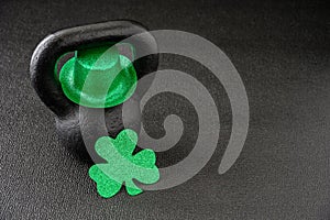 Black iron kettlebell and green glitter shamrock and leprechaun hat on a black gym floor, happy St. Patrick’s Day