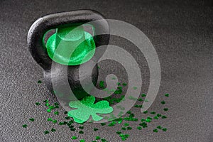 Black iron kettlebell, and green glitter shamrock and leprechaun hat, covered in green shamrock confetti on a black gym floor, hap