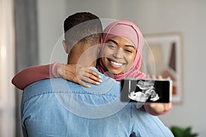 Black Islamic Lady Embracing Husband And Holding Smartphone With Baby Sonography Photo