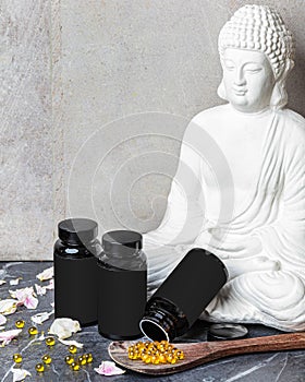 Black jar with yellow oil capsules in wooden spoon ower white buddha figurine and flower petals