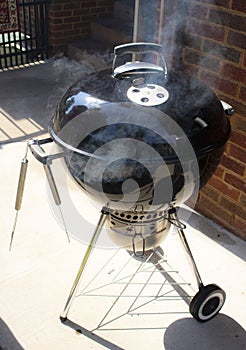 Black kettle type outdoor charcoal grill sitting on a patio beside a brick wall with smoke coming out of it and a pair on tongs ha