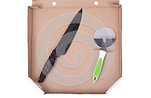 A black kitchen knife and a pizza cutter are in an open cardboard pizza box, isolated on white, top view