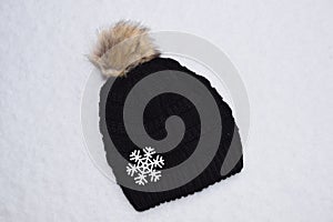 Black hat with furry ball and large snowflake on fresh snow.